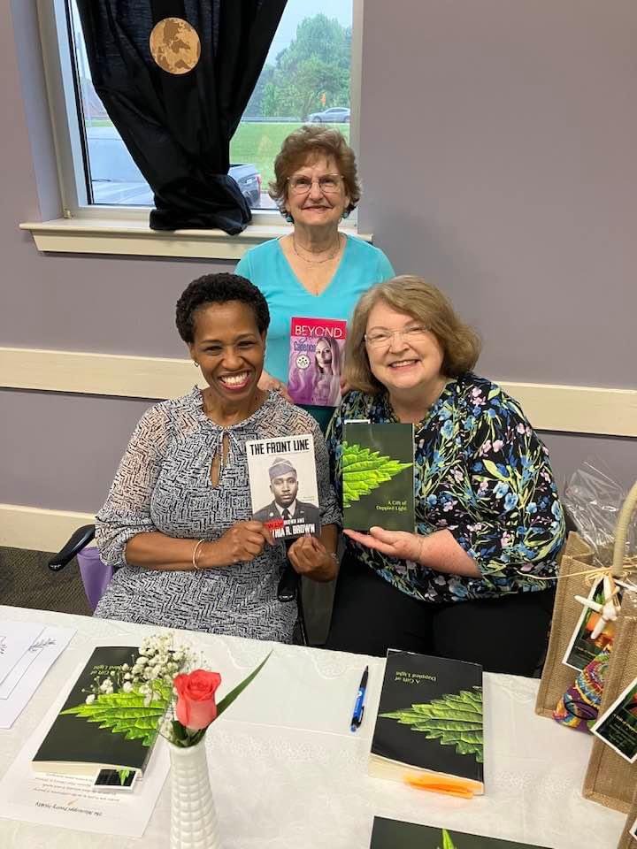 Three people posing with books at a table displaying titles and flowers.