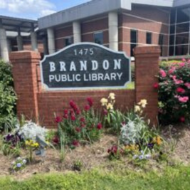 Brandon Public Library sign with flowers in front and red brick building in the background.