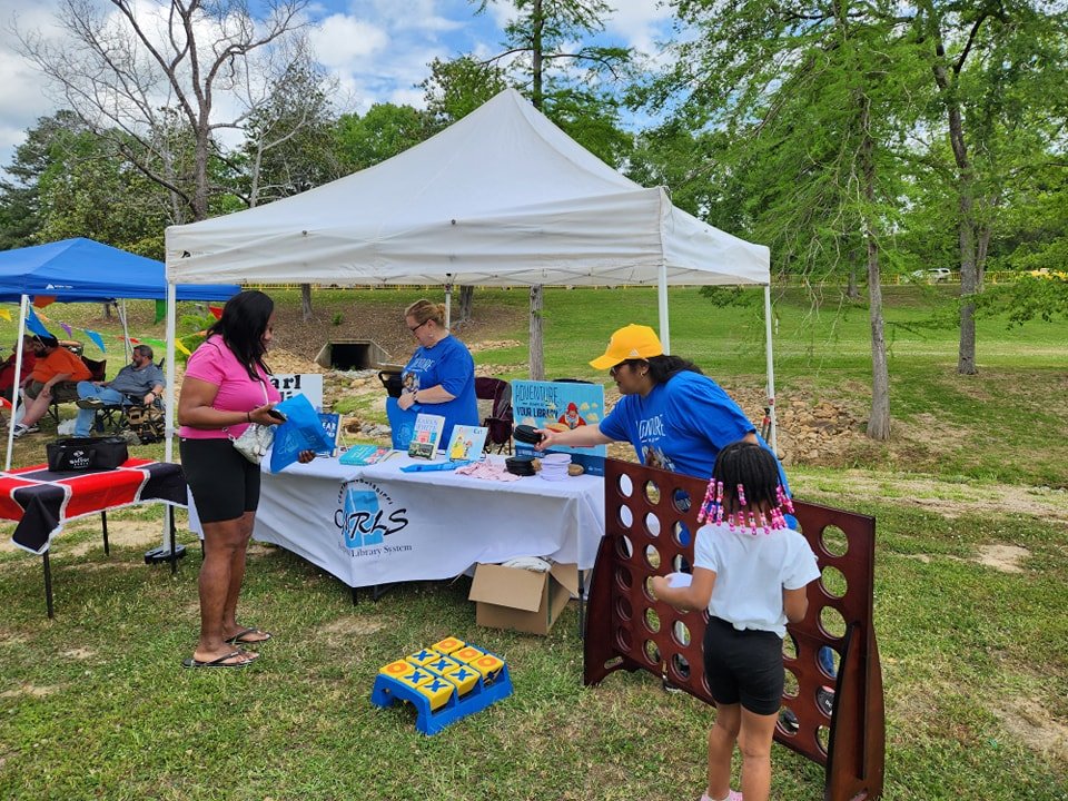 Outdoor literacy event with a tent, table displaying books, people interacting, and a child playing Connect Four.