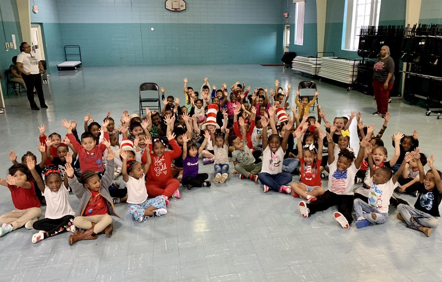 Group of children sitting on a gym floor with hands raised, some wearing red and white striped hats.