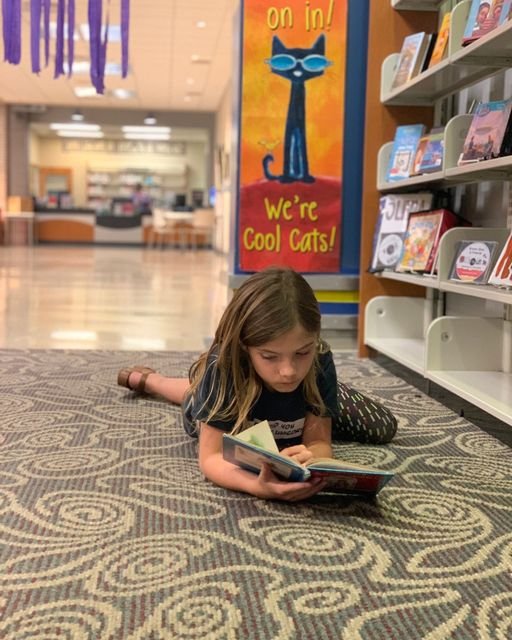 Child lying on the floor reading a book in front of a bookshelf, with a poster that says "We're Cool Cats!" nearby.