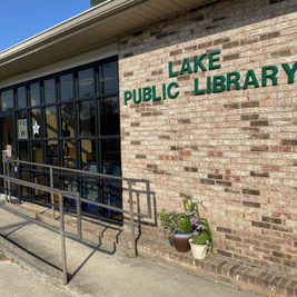 Entrance of Lake Public Library with glass doors, brick wall sign, and metal accessibility ramp.