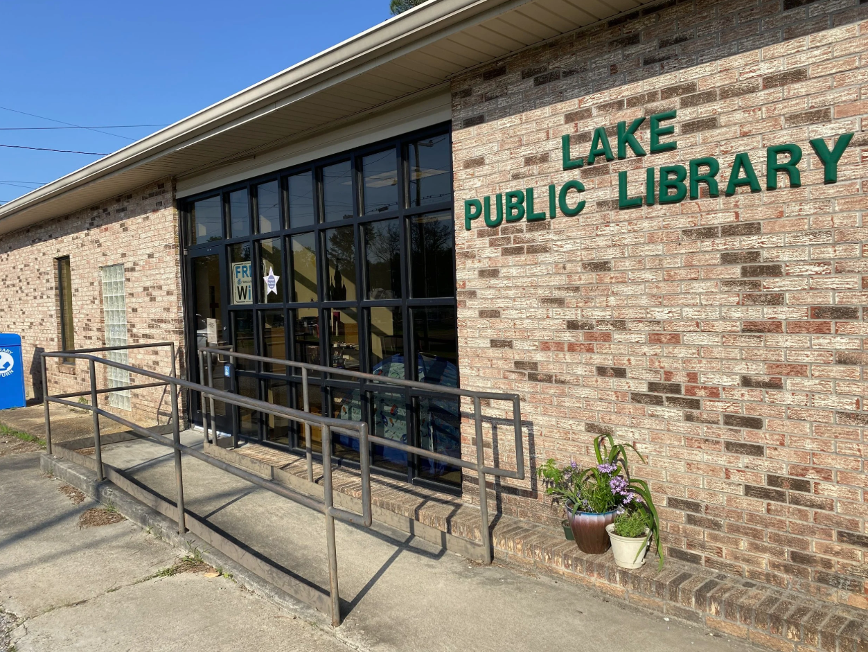 Entrance of Lake Public Library with glass doors, brick wall sign, and metal accessibility ramp.