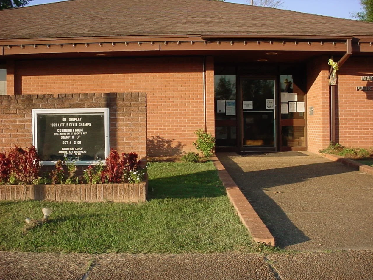 Mendenhall Public Library with red brick exterior, glass entrance, and event sign near walkway.