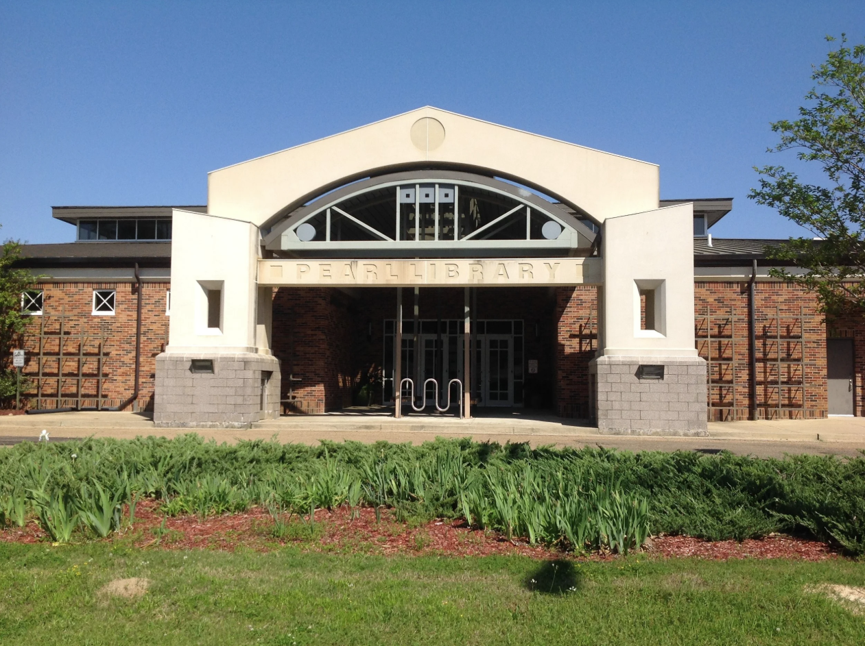 Front entrance of Pearl Public Library featuring a large arched design, glass doors, brick walls, and landscaped greenery in the foreground.