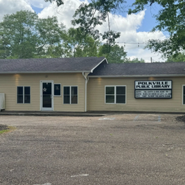 Polkville Public Library with yellow facade, book return box, and hours sign near entrance.