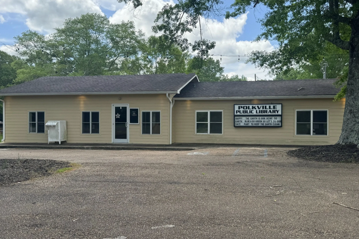 Polkville Public Library with yellow facade, book return box, and hours sign near entrance.