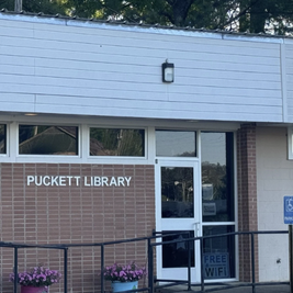 Entrance of Puckett Public Library with brick facade, white door, "PUCKETT LIBRARY" sign, potted flowers, and a "FREE WIFI" notice on the door.