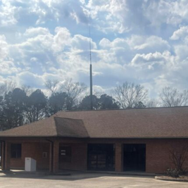 Brick building with gabled roof and tall antenna, surrounded by trees under a partly cloudy sky.