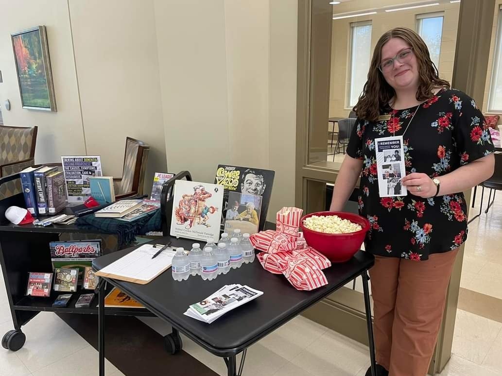 Miranda standing next to a table with popcorn, water bottles, and pamphlets, beside a cart filled with books and DVDs.