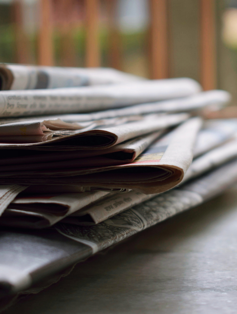 Stack of folded newspapers placed on a flat surface with soft natural light coming through wooden bars in the background.