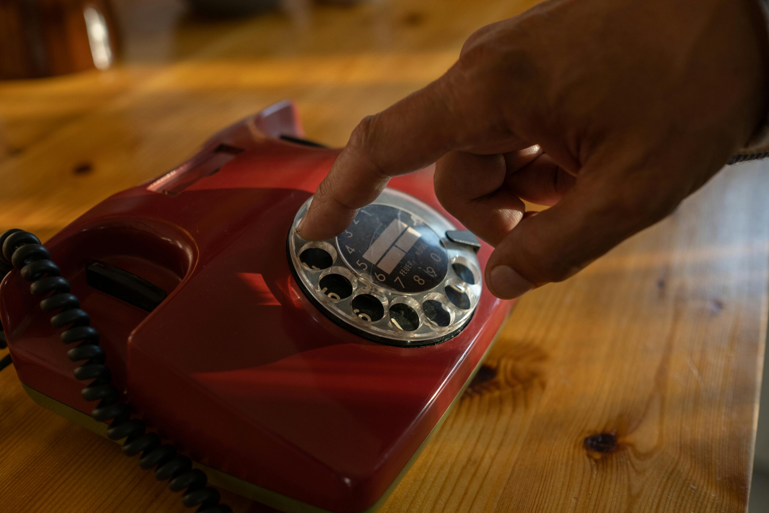 Hand dialing a number on a red rotary phone placed on a wooden table.